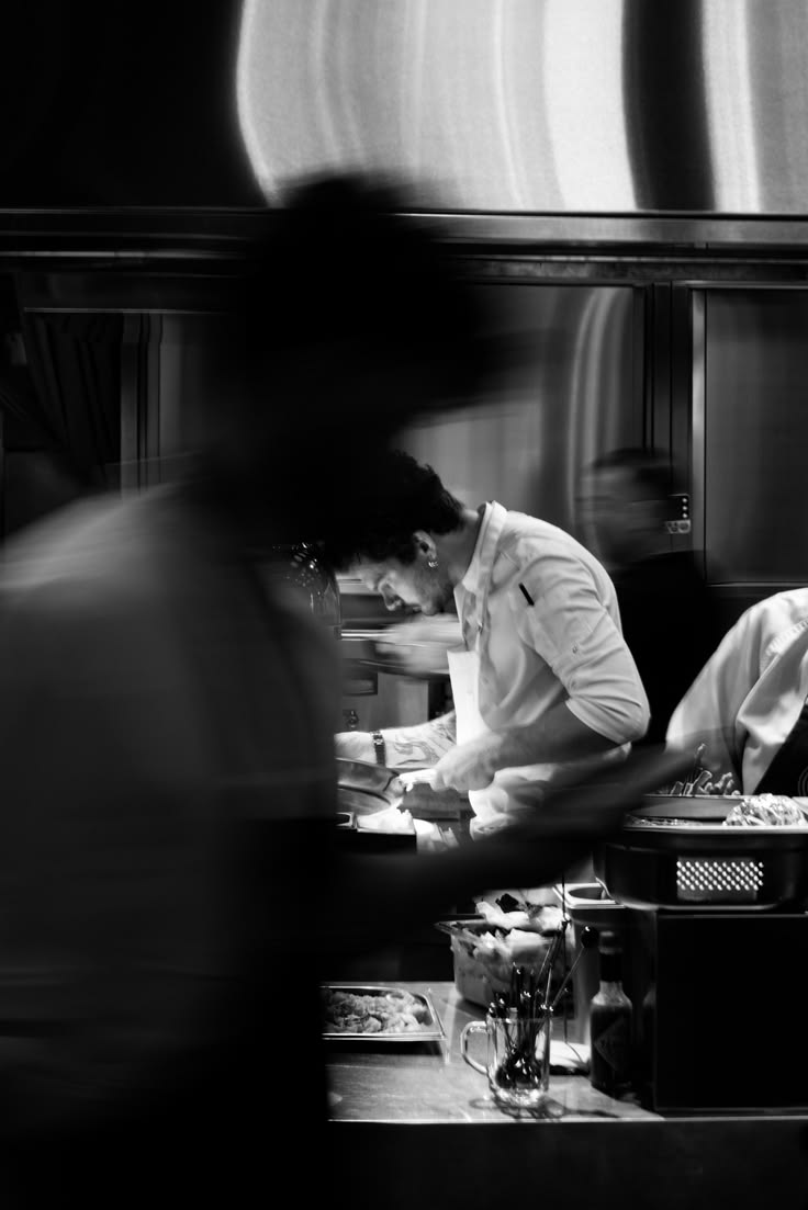 A chef at work in a restaurant kitchen, seen behind the blurred motion of a passing colleague.